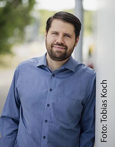 Ein Mann in Hemd und Hose steht lächelnd vor einem Gebäude.