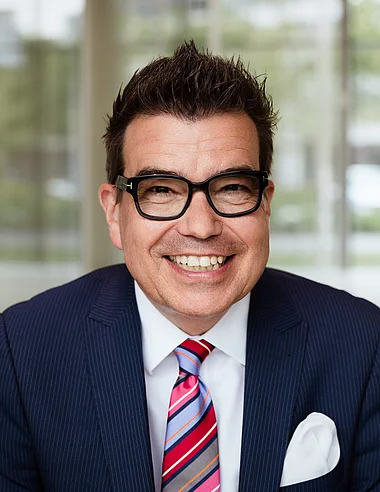 Ein Mann im Anzug lächelt professionell vor einem modernen Hintergrund.